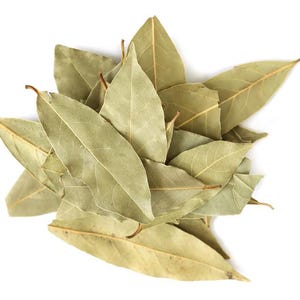 May include: A cluster of dried bay leaves, each featuring an elongated, oval shape with a pointed tip. The leaves exhibit a muted, pale olive-green color with visible central veins and a smooth, matte texture, arranged in a layered, overlapping pile.