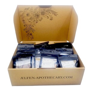May include: A cardboard box with the top open, filled with small, sealed packets. The box has a floral design on the inside of the lid and the text "ÆLFEN-APOTHECARY.COM" on the front. The packets are black and white.