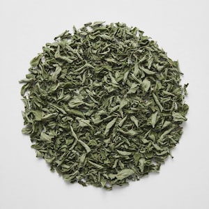 May include: A circular arrangement of dried, dark green mint leaves. The leaves are crinkled, fragmented, and varied in size, creating a textured, organic pattern against a plain, neutral, light-colored background surface.