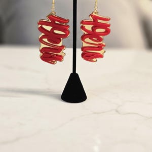 May include: A pair of gold-toned dangle earrings with a red cursive word design. The earrings are displayed on a black stand against a blurred background. The word design is repeated on each earring.
