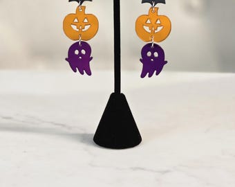 May include: A pair of Halloween-themed dangle earrings. Each earring features a black bat charm, an orange pumpkin charm, and a purple ghost charm. The top of each earring is a black and orange beaded ball.