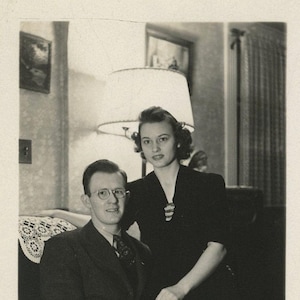 May include: Black and white vintage photograph of a man and woman. The man wears glasses, a suit, and tie, seated on a couch. The woman stands behind him in a dark dress, with a brooch. A lamp and artwork are visible.