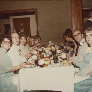 May include: A group of people seated at a long dining table covered with a white tablecloth, set with plates, glassware, and food. The individuals are wearing formal attire, including light blue dresses and suits, in a vintage indoor setting.