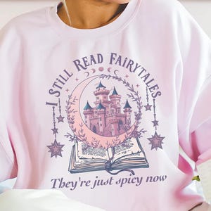 Op de afbeelding: Lichtroze sweatshirt met de tekst "I STILL READ FAIRYTALES" en "They're just spicy now". De afbeelding bevat een kasteel, maan, sterren en een open boek. Dit sweatshirt maakt deel uit van een kledingcollectie.