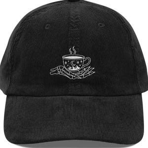 Könnte beinhalten: Schwarze Cord-Baseballkappe mit weißem Stickdesign. Das Design zeigt eine Kaffeetasse mit einer Berg- und Mondszene, die von einer Skeletthand gehalten wird. Die Kappe hat einen gebogenen Schirm und einen Knopf oben.