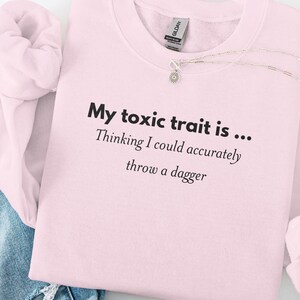 May include: A light pink sweatshirt with the text "My toxic trait is... Thinking I could accurately throw a dagger" in black. The sweatshirt is paired with ripped blue jeans and a silver necklace.
