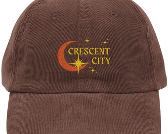 May include: A brown corduroy baseball cap with the text "CRESCENT CITY" in yellow and an orange crescent moon and star design. The cap has a curved peak and a fabric strap.