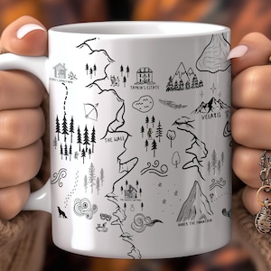 Może przedstawiać: Biały ceramiczny kubek z czarno-białym wzorem mapy, zawierającym ilustracje gór, drzew i budynków. Kubek trzyma osoba w beżowym swetrze.