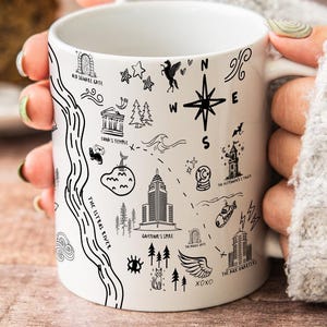 A person's hands holding a white mug with various black and white illustrations and text on it, placed on a wooden surface.
