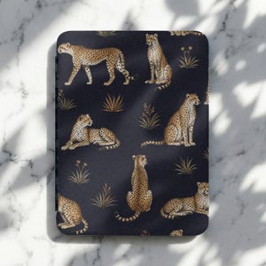 May include: A rectangular e-reader cover featuring a dark navy blue background with a repeating pattern of cheetahs in various poses, including sitting, lying down, and walking, interspersed with small golden-brown tufts of grass and plants.