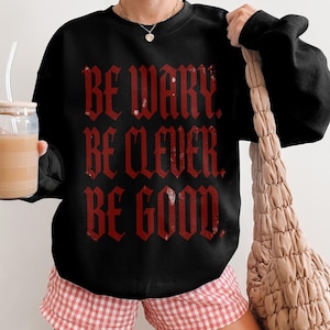 Black sweatshirt with red gothic text "BE WARY. BE CLEVER. BE GOOD."