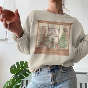 May include: Beige sweatshirt with a holiday-themed graphic. The graphic depicts a bookstore window with books, a Christmas tree, and the text "READ FOR THE HOLIDAYS." The sweatshirt is paired with blue jeans.