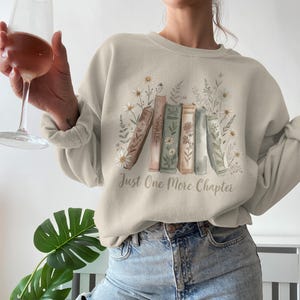 May include: Beige sweatshirt with a floral and book design. The graphic features a row of books with floral accents and the text "Just One More Chapter". The sweatshirt is a neutral color and has long sleeves.