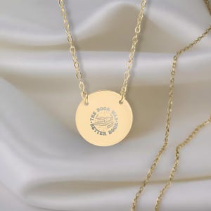 May include: Gold-colored necklace with a circular pendant. The pendant has the engraved text "THE BOOK WAS BETTER BOOK" around a stack of books. The chain is delicate and the background is a soft, white fabric.