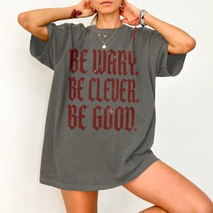 Dark grey t-shirt with bold red text "BE WARY. BE CLEVER. BE GOOD."