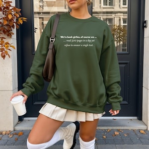 May include: An olive green sweatshirt with the text "We're book girlies, of course we... read 500 pages in a day yet refuse to answer a single text." The sweatshirt is paired with a white pleated skirt and white boots. A brown purse is worn over the shoulder.