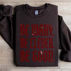 Brown sweatshirt with red text "BE WARY. BE CLEVER. BE GOOD."