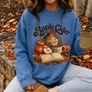 Blue sweatshirt with "Apple Cider a crisp autumn classic" graphic and autumn elements.