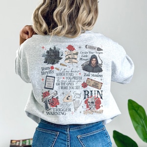 May include: A light gray sweatshirt featuring various graphic designs and text. The designs include a castle, a person, a gun, a skull, and roses. Text includes "Run", "Eyes on Me", "Trigger Warning", and "Little Mouse".