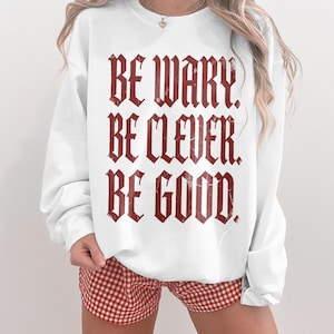 White crewneck sweatshirt with "BE WARY. BE CLEVER. BE GOOD." in gothic font.
