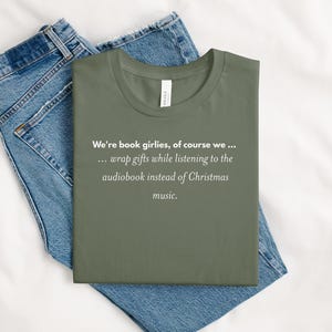 May include: Olive green t-shirt with the text "We're book girlies, of course we... wrap gifts while listening to the audiobook instead of Christmas music." The shirt is folded on top of blue jeans.