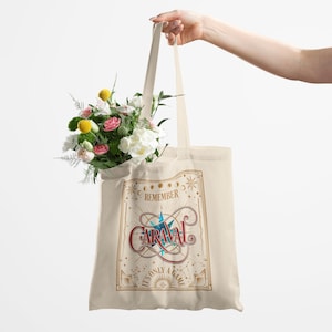 May include: Cream-colored tote bag with the text "Remember Caraval, It's Only a Game" in a decorative design. The bag is filled with a bouquet of flowers in shades of pink, white, and yellow. The bag is held by a person.