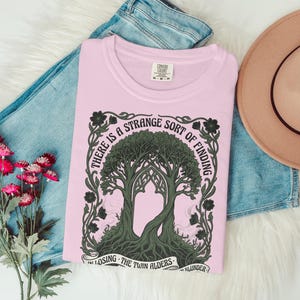 Pink t-shirt with tree design and text "There is a strange sort of finding in losing the twin alders."