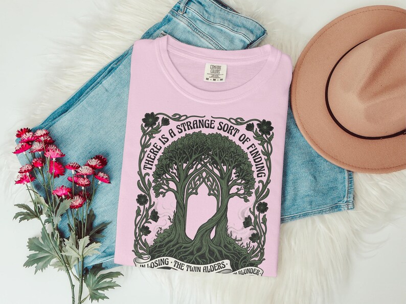 Pink t-shirt with tree design and text "There is a strange sort of finding in losing the twin alders."