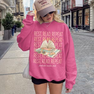 Pink sweatshirt with "REST, READ, REPEAT" design featuring an open book.