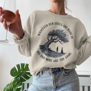 Może przedstawiać: Beżowa bluza z nadrukiem drzewa i dwóch postaci, z napisem "Whatever our souls are made of, his and mine are the same." Bluza jest połączona z niebieskimi dżinsami.