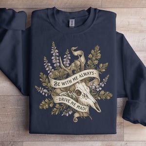 Może przedstawiać: Granatowa bluza z nadrukiem przedstawiającym czaszkę jelenia, poroże i elementy kwiatowe. Na banerze widnieje napis "BE WITH ME ALWAYS - DRIVE ME MAD!". Projekt w odcieniach kremu, brązu i zieleni.