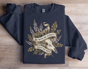 May include: Navy blue sweatshirt featuring a detailed graphic of a deer skull, antlers, and floral elements. A banner across the skull reads "BE WITH ME ALWAYS - DRIVE ME MAD!" The design is in shades of cream, brown, and green.