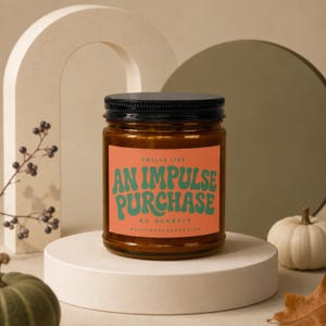 May include: A brown glass candle jar with a black lid and an orange label. The label features green text that reads: SMELLS LIKE AN IMPULSE PURCHASE NO REGRETS MAGICMERCHEMPORIUM. The candle is centered on a circular white platform.