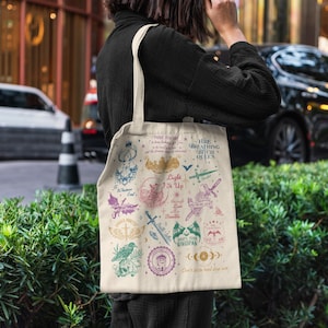 Puede incluir: Bolso tote beige con ilustraciones y texto coloridos. El diseño incluye varios símbolos, espadas y las palabras "Here Breathing Witch Queens". El bolso se lleva al aire libre.