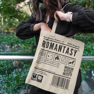 Op de afbeelding: Beige draagtas met het woord "ROMANTASY" in dikke zwarte letters. De tas heeft verschillende pictogrammen en tekst, waaronder "100% Organic" en een barcode. De tas wordt vastgehouden door een persoon die een zwarte top draagt.
