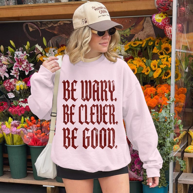 Light pink sweatshirt with "BE WARY. BE CLEVER. BE GOOD." in gothic font.