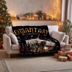 May include: A navy blue blanket with gold lettering reading "ROMANTASY READERS SOCIETY" is draped over a white sofa. The blanket design features illustrations of books, roses, and butterflies. The phrase "JUST ONE MORE CHAPTER" is also visible.