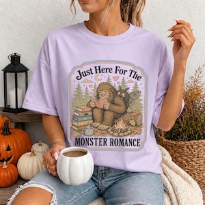 May include: A light purple t-shirt featuring a graphic print of a brown sasquatch reading a book next to a moth and a campfire. The text reads Just Here For The MONSTER ROMANCE. The design includes pine trees, hearts, and a stack of books.
