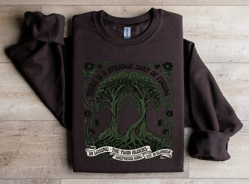 Dark brown sweatshirt with green tree design and text: "There is a strange sort of finding in losing the twin alders. Shepherd King est. in blunder."