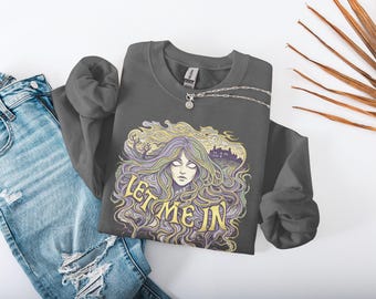 May include: Dark gray sweatshirt with a graphic of a woman with long, flowing hair and the text "LET ME IN". The design features shades of purple, yellow, and green. A silver necklace and blue jeans are also in the image.