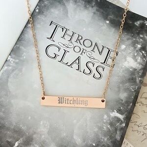 A gold-colored necklace with a rectangular pendant engraved with the word "Witchling" is displayed on top of a book titled "Throne of Glass".