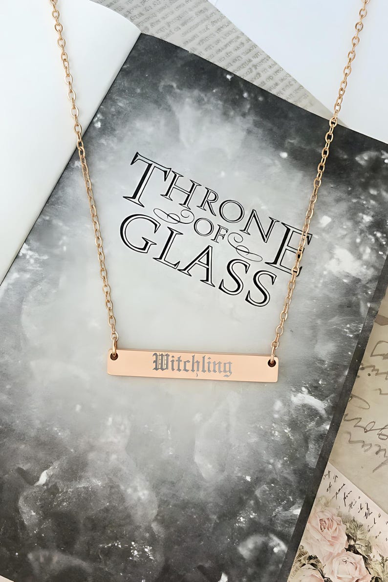 A gold-colored necklace with a rectangular pendant engraved with the word "Witchling" is displayed on top of a book titled "Throne of Glass".