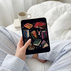 May include: Black tablet case featuring a colorful illustration of open books with gold star accents. The case has a black background and is held by a person wearing blue and white striped pajamas.