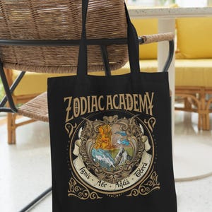 May include: Black tote bag featuring the words "ZODIAC ACADEMY" in gold lettering. The bag displays a detailed crest with fire, water, air, and earth symbols. The bag has black handles and is hanging on a chair.