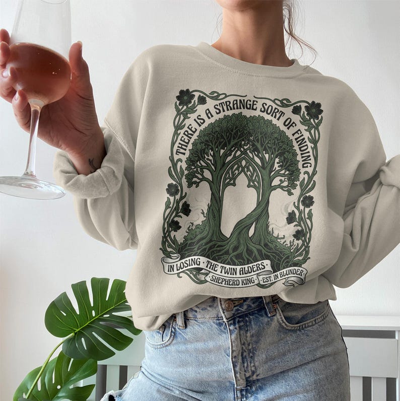 Sweatshirt with twin alder trees and text "There is a strange sort of finding in losing the twin alders"