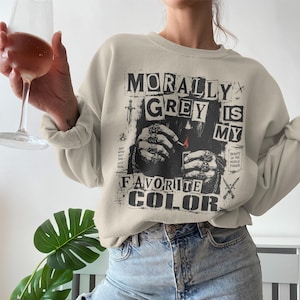 Może przedstawiać: Beżowa bluza z napisem "MORALLY GREY IS MY FAVORITE COLOR" w kolorze czarnym. Grafika zawiera dłoń trzymającą zapalniczkę i inne gotyckie obrazy. Bluza jest połączona z niebieskimi dżinsami.