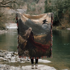May include: A woven tapestry blanket featuring a fantasy landscape with a woman in a red cloak and a dragon. The blanket has a fringed edge with shades of red, brown, and black. The scene depicts a mountain range and a river.