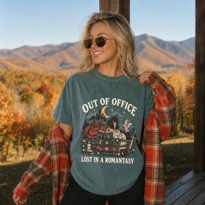 Peut inclure: Un t-shirt bleu ardoise à manches courtes avec une impression graphique représentant une personne lisant sur une pile de livres avec un dragon, une épée et un croissant de lune. Le texte indique "OUT OF OFFICE" et "LOST IN A ROMANTASY".