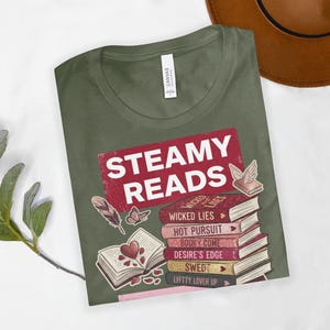 May include: Olive green t-shirt with the text "STEAMY READS" in a red rectangle. Below the text is a stack of books with titles like "Wicked Lies" and "Hot Pursuit". Decorative elements include a feather, butterfly, and open book.