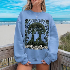 Light blue sweatshirt with ornate tree design and text "There is a strange sort of finding in losing the twin alders."